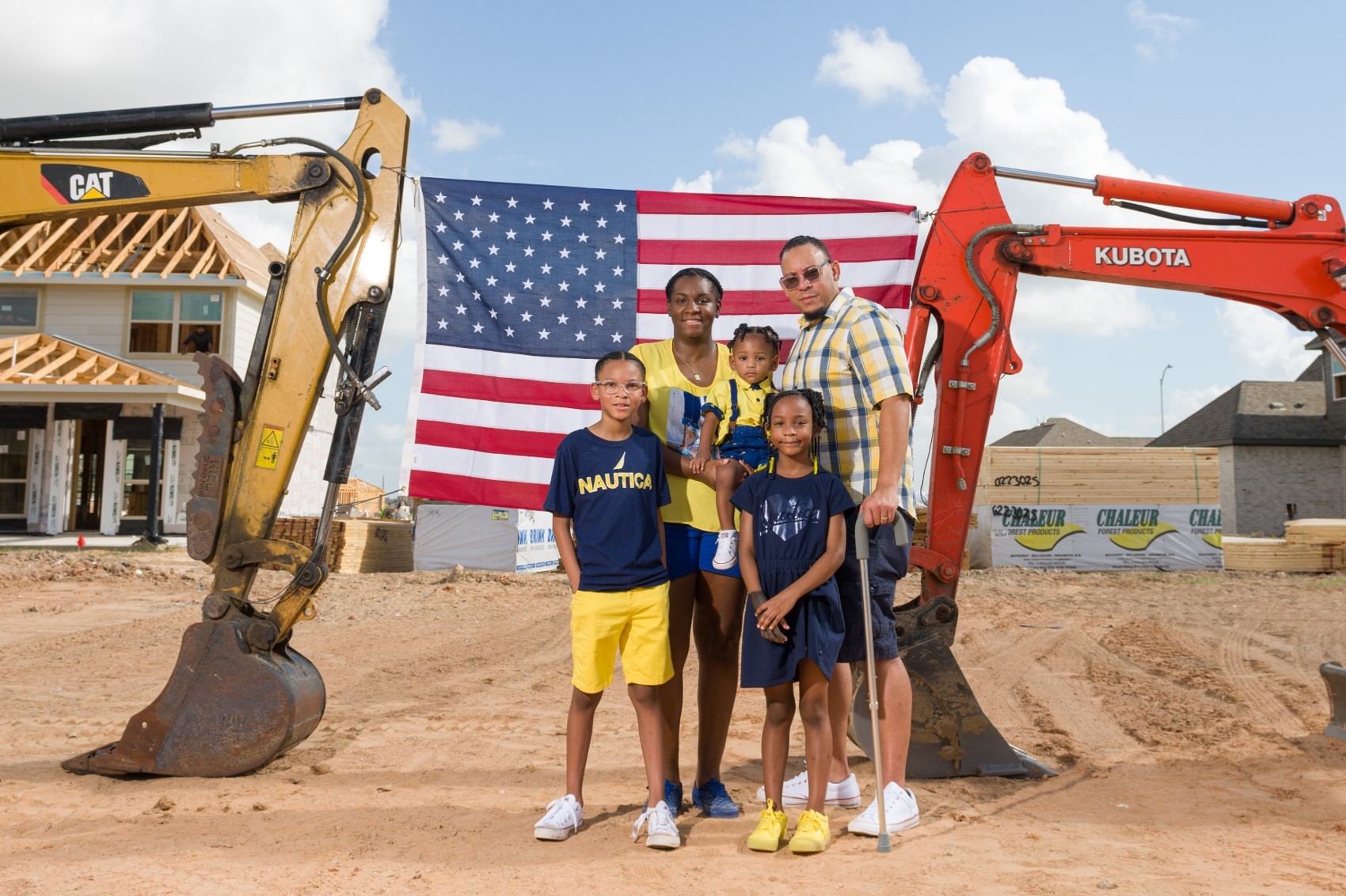 Family standing in front of the construction site of their home. Two excavators flank them with an American flag spread between them in the background.